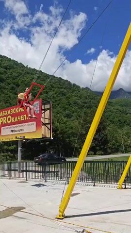 The video features a giant swing ride set in a mountainous region of Bzypta, Abkhazia, on a bright, sunny day with a clear blue sky and scattered white clouds.

The main subjects are two people, appearing to be adults, seated side-by-side in a red swing harness. The ride structure includes two prominent tall, bright yellow metal poles and a red launching mechanism at the base. At the beginning of the video (0:00-0:01), the swing is seen held by the red structure on a concrete platform, with an operator seemingly preparing for launch. A red and white banner is partially visible in the background, showing some Cyrillic text, including "смелых" (brave/daring).

From 0:02 onwards, the swing is released, propelling the riders high into the air. The perspective shifts to show the swing soaring over a wide, fast-flowing river with light brown/grey water. The background is dominated by lush, green, heavily forested mountains that rise steeply from the riverbanks. The activity clearly depicts a thrill ride, with the swing reaching a significant height and arc, suggesting an exhilarating experience for the participants.

The setting is an outdoor adventure park or attraction, leveraging the dramatic natural scenery of Abkhazia, including the mountains and river. The time of day is daytime, indicated by the bright sunlight and clear skies.
