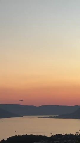 The video shows an airplane flying over a body of water at sunset. The sky is a gradient of orange and pink. The airplane is a silhouette against the colorful sky. In the foreground, there are hills and trees.