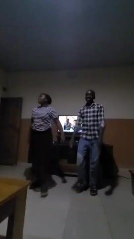 Two young people, a girl and a boy, are dancing in front of a television screen. The girl is wearing a striped t-shirt and a long black skirt, while the boy is dressed in a plaid shirt and jeans. The room appears to be a living room or common area, with a television set on a stand, a wooden door, and a table visible in the foreground. The lighting suggests it is either nighttime or a dimly lit indoor space. The overall mood seems energetic and joyful, as indicated by their dance moves and smiles. The TV screen displays an image, possibly from a show or video they are dancing to. The scene is set in Kuru Karama, Nigeria.