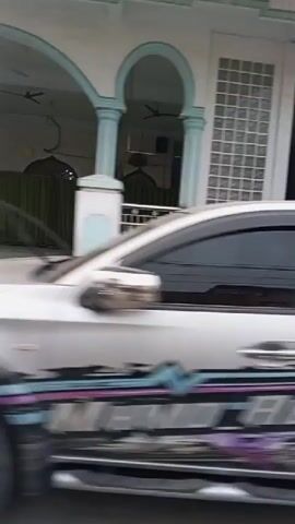 The video begins with a close-up, blurred shot of a white pickup truck with a complex graphic design on its side. The truck is moving, as indicated by the motion blur. The scene then shifts to a wider view of an outdoor area, possibly a street or driveway, with buildings on either side. One building appears to have a large, multi-panel garage door. Another building is white and has windows.

The video continues to show a woman wearing a black hijab and an orange top walking past a red metal gate. She is slightly blurred. Next, the perspective changes again to show a street scene with a white building on the left. On the right, there is a small food or drink stall with an umbrella and a display case. A person is seated behind the stall. The scene is also blurred due to movement. The video continues to pan across this area before abruptly ending.