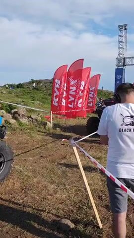 This media file captures an outdoor event, likely a race or competition, with an all-terrain vehicle (ATV) as a central object. The scene is set on a dry, grassy terrain, suggesting a rural or off-road location. The time of day appears to be afternoon, indicated by the bright sunlight and the presence of shadows. The weather is fair, with a partly cloudy sky.

In the foreground, a blue and black ATV, heavily coated in mud, is positioned on the ground. Its large, knobby tires and rugged construction suggest it is built for challenging terrains. In front of the ATV, a person wearing a white t-shirt with black lettering is standing, facing away from the camera. The text on their shirt reads "BLACK FLIGHT." Adjacent to them, a red and white striped ribbon marks a boundary or course.

In the background, a large start/finish gate structure is visible, adorned with various logos and text. Prominently displayed are several tall, red flags with white lettering that reads "LYNX." These flags, along with the gate, imply an organized event. Several other individuals are present, some appearing to be participants or staff. One person in an orange vest is standing near the gate, possibly overseeing the event. Another individual in a dark jacket is standing further back, looking towards the gate. The overall atmosphere seems to be one of anticipation and preparation for an event.

The context provided indicates this scene is located in Antarut, Armenia. This geographical information, combined with the visual cues of the landscape and event setup, paints a picture of an off-road racing event taking place in the Armenian countryside.