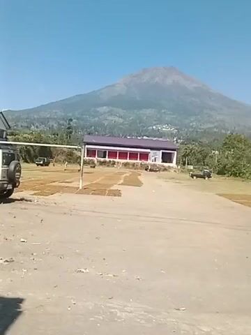 The video begins with a wide shot of a flat, dusty area with a building and a large mountain in the background. The building appears to be a single-story structure with a red trim and a dark roof. In front of the building, there are piles of what looks like dried leaves or grass. A white SUV is parked to the left of the frame. As the video progresses, a person on a motorcycle wearing a hooded jacket rides into the frame from the left. The motorcycle is black and appears to be a scooter-style bike. The person on the motorcycle is wearing a dark helmet. The camera continues to pan slightly, showing more of the dusty area and the mountain in the distance. A few other vehicles, including a dark-colored truck, are visible in the background. The sky is clear and blue. The video ends with a similar shot to the beginning, focusing on the building and the mountain.