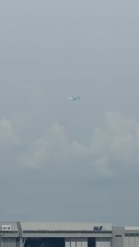 A wide shot shows an airplane flying through a cloudy sky. The airplane is small in the frame and appears to be light blue. Below the sky is the top of a building, possibly an airport terminal. The building is white and has large windows and a gray roof. The word "ANA" is visible in blue lettering on the side of the building. The sky is overcast with numerous gray clouds.