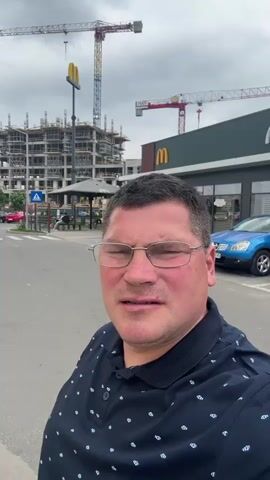 The video is a close-up selfie shot of a middle-aged man with short graying hair, wearing glasses and a dark blue collared shirt with white polka dots. He is standing on a street. Behind him is a McDonald's restaurant with a McCafe section. A blue car is parked to the side of the McDonald's. Across the street is a tall, unfinished building with construction cranes. The sky is overcast. The man is looking at the camera and talking.