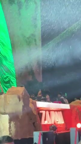 A crowd is gathered around a stage with the words "ZANNA" illuminated on the front. A DJ is performing behind the nameplate. There is a giant green structure behind them and above the stage a mist hangs in the air. People are taking pictures and videos with their phones. Some people have their hands in the air. The environment is dark, lit by stage lights and the glow of the sign.
