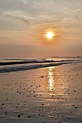The video captures a serene beach scene at sunset. The sun, a bright orb, sits low in the sky, partially obscured by a thin layer of clouds.  Its golden light reflects off the wet sand of the beach, creating a bright path towards the viewer. Small, gentle waves roll in from the sea, their crests just visible in the distance. The beach is covered with small pebbles, which add texture to the foreground. In the far distance, the land can be seen as a dark silhouette against the light of the setting sun. The sky is a mix of pale blues and oranges, streaked with light clouds.