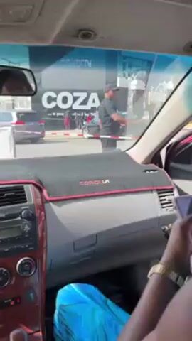 The video shows the interior of a car, likely a Toyota Corolla, with a red dashboard and door panels.  A person wearing a blue, green, and black patterned top is sitting in the back seat and opens the car door. Another person, wearing a white floral top and maroon pants, exits the car after the first person. They are wearing pink Crocs.  Outside the car, a man in a black shirt and hat stands near a barrier with a large sign that reads "COZA" visible in the background.  Other cars and people are also visible outside the car.