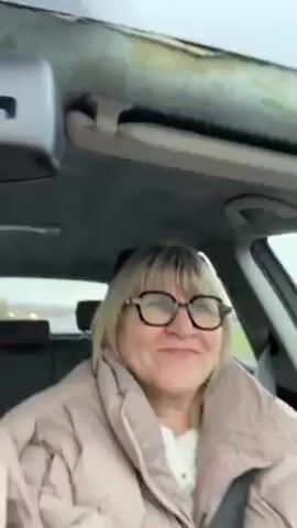 A woman wearing glasses and a tan puffer jacket is sitting in the driver's seat of a car. She looks directly at the camera and opens her mouth slightly. The car appears to be in motion, with a road visible in the background. The interior of the car shows wear and tear, particularly on the ceiling.