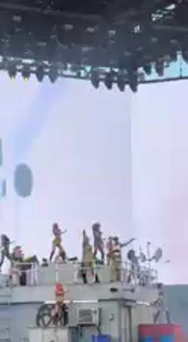 Multiple performers are visible on a large outdoor stage during what appears to be a concert or festival. The stage is equipped with overhead lighting rigs and a massive LED screen in the background displaying dynamic blue and white light patterns, resembling abstract energy bursts or clouds. A large, disc-shaped object, possibly a speaker or decorative element, hangs on the left side of the stage. The performers are actively moving and gesturing, indicative of a live musical or dance performance. The bright lighting suggests it could be daytime, possibly late afternoon or early evening. The scene conveys an energetic atmosphere typical of a large-scale event, set in Indio, United States.
