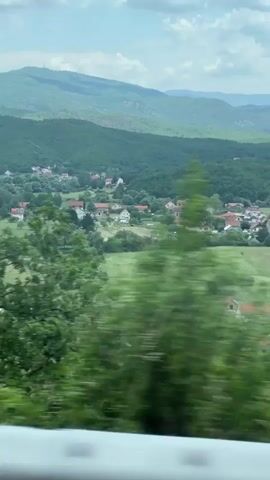The video shows a countryside view from a moving vehicle, likely a train or bus. The landscape is characterized by green fields and houses nestled amongst trees. In the background are rolling hills and a distant mountain range. A large red and yellow building is visible at one point. The sky is partly cloudy.