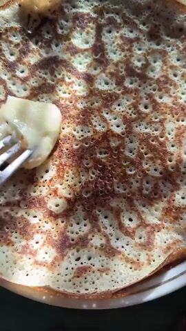A close-up shot shows a fork with a piece of butter on it hovering over a stack of golden-brown pancakes. The pancakes have a bubbly texture and appear to be freshly made. Sunlight illuminates the scene, highlighting the deliciousness of the food.