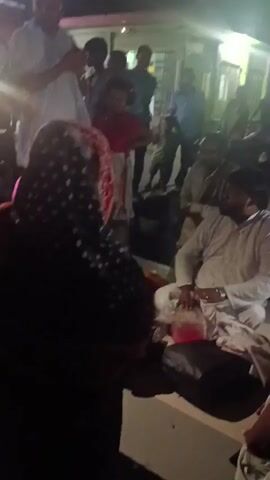 A group of people are gathered at night, illuminated by artificial lights. In the foreground, a woman wearing a dark headscarf and patterned shawl is visible from behind. She appears to be interacting with someone or something out of frame. To her right, a man in a white outfit is seated on a dark surface, holding a clear bag with pink liquid. Behind them, a child in a red top and a patterned skirt stands facing away from the camera. Other individuals are standing and milling about in the background. The environment appears to be outdoors, possibly a street or an open area, with a building visible in the background. Some people are holding mobile phones, suggesting they might be recording or using their devices. The overall scene suggests a gathering or an event taking place in a public space at night.