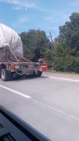 A truck is seen driving on a highway. The truck is carrying a large, white, tarp-covered load. The truck has multiple axles and large tires. The video is shot from inside another vehicle, presumably a car, looking out the window. The surrounding environment appears to be rural, with trees and bushes visible alongside the road. The sky is blue with some scattered clouds. The truck appears to be in motion, and the video has a slight blur, indicating movement.