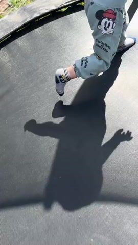 A close-up shot shows a toddler's legs as they stand and move on a black trampoline. The toddler is wearing light green pants with a repeating pattern of Mickey Mouse characters, and a white t-shirt with black markings. Their feet are clad in light-colored shoes with blue accents. The toddler's shadow is cast prominently on the trampoline surface. The trampoline mat appears worn in places, and its edge, a black band with yellow springs, is visible. The background is blurred, showing hints of green grass and sunlight. The camera angle is from above, focusing on the toddler's lower body and the trampoline surface.