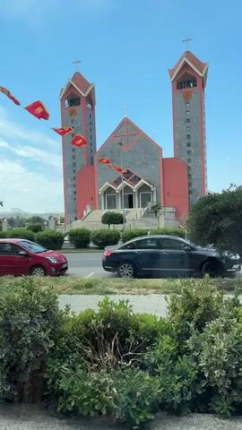 The video displays a modern church under a clear blue sky. The church features a distinctive design with two tall, rectangular bell towers on either side, each topped with a cross and a clock face. The facade is a blend of grey stone and reddish-orange panels, with a large cross adorning the central structure above the main entrance. Red flags, bearing a golden emblem, are strung across the street in front of the church, adding a decorative element to the scene. In the foreground, lush green bushes line the edge of a road where a red hatchback and a black sedan are parked. The bright lighting suggests it is daytime with sunny weather.