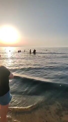 The video shows a beach scene at sunset. The sun is low in the sky, casting a warm glow. There are several people in the water, swimming and playing. The water is calm, with gentle waves lapping at the shore. In the foreground, a person stands on the beach, facing the water. The overall atmosphere is peaceful and relaxing.
