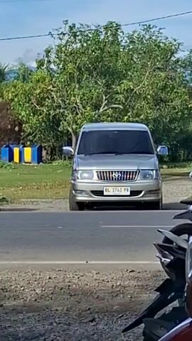 The video shows a silver Toyota Kijang Innova parked on the side of a road. A scooter is passing by the car from right to left. The car has a license plate that reads BL 1761 PV. In the background, there are trees, some blue and yellow barriers, and a blue sky with some clouds. The road is paved and has a gravel shoulder.