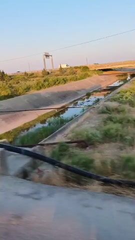 The video shows a view from a moving vehicle. The scene includes a concrete canal with water flowing in it. The canal is flanked by grassy slopes with some vegetation. In the background, there is a road with a truck driving on it. The sky is clear and blue with the sun setting, casting a warm light on the landscape. The camera pans to the right, revealing more of the natural surroundings, including trees and shrubs.