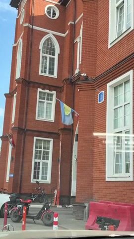 The video shows a building with a red brick facade. It has white-framed windows, and a number "1" is visible on the corner of the building. A flag of sorts hangs on the building. It is a light blue fabric with a yellow circle featuring a face design in the center. The flag is partially obscured and appears to be slightly tattered. Two scooters are parked in front of the building. A white car is parked behind the scooters.