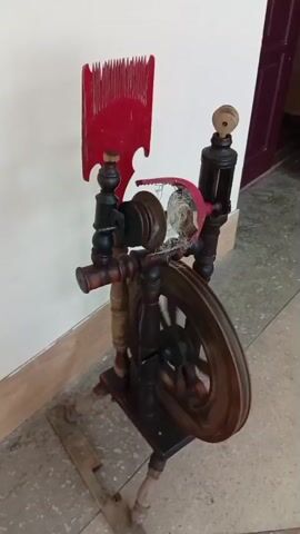 The video shows a close-up view of a spinning wheel. The spinning wheel is made of dark wood and has a large wheel with spokes. There is a spindle and flyer mechanism for spinning yarn. A red comb-like tool is attached to the top of the spinning wheel. The spinning wheel is positioned against a light-colored wall, and the floor is tiled. The video pans and zooms in on different parts of the spinning wheel, highlighting its intricate details and craftsmanship.