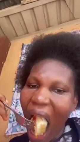 A woman with short, dark curly hair is looking into the camera as she eats food from a fork. She is wearing a floral shirt. Behind her is a window with a patterned curtain. The room is partially visible with a brown piece of furniture and a corrugated white ceiling. The woman brings the fork to her mouth, chews and swallows the food, and then looks directly at the camera again.