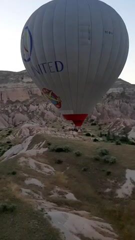 A wide shot shows a hot air balloon with the word "UNITED" and a logo on its side. The balloon is gray with blue lettering and a colorful graphic. Below the balloon, a red basket is visible, with people inside. The balloon is in the air over a landscape of rolling hills and rock formations. The sky is clear and bright. The video is approximately 4 seconds long.