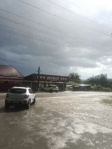 This video shows a street scene during a heavy rain shower in Ivanovskaya, Russia. The sky is overcast with dark storm clouds, and rain is visibly falling, creating ripples on the wet asphalt and splashing as it hits. The ground is covered in puddles.

In the foreground, a white car is parked on the left side of the road. Further down the street, another white car and a dark car are visible. A building with a sign displaying Cyrillic text is seen on the right side of the street, behind a fence. The text on the sign appears to read "БУРЖУЙ" with smaller text below it. A utility pole with power lines runs across the top of the frame. The overall mood is one of a sudden downpour, with the weather being the most dominant element. The time of day appears to be daytime, given the brightness of the sky despite the clouds.