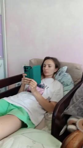 A woman is sitting in a chair, holding a green phone in her hands. She is wearing a white t-shirt and green shorts. Her hair is brown and she is looking at the phone. The background is a light pink wall. The video is 5 seconds long.