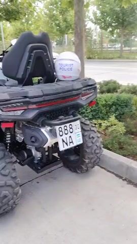 The video shows a close-up, slightly shaky, exterior view of a grey and black all-terrain vehicle (ATV) parked outdoors. The ATV is facing towards the right side of the frame, with its rear end prominently displayed at the beginning of the video. The license plate on the rear of the ATV reads "888 NA UZ". A white helmet with "TOURIST POLICE" written on it in blue is secured to the back of the ATV's seat. The ATV has large, knobby tires and a robust suspension system. In the background, a white car is visible, and trees and bushes line the area. The ground appears to be paved. The camera pans slowly from the rear of the ATV towards the front, showing more of its design and features.