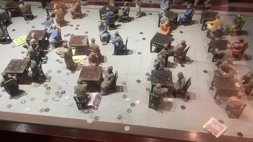 The video shows a diorama depicting a courtyard with miniature figures sitting at tables. The figures are arranged in groups around the tables, giving the impression of people conversing or playing games. Small, silver coins are scattered on the ground around the tables. The diorama is enclosed in a glass case.