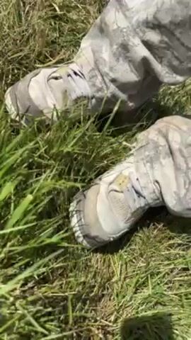 Here's a detailed description of the video:

The video starts with a close-up of a person's legs from the knees down. The individual is wearing camouflage-patterned pants and light-colored, worn sneakers. They are standing on a patch of green grass. 

The camera then pans up to reveal the full outfit of the person. They are wearing camouflage pants, a sleeveless gray t-shirt, and a dark green cap. The person is standing in what appears to be a campsite or festival grounds, with numerous tents visible in the background. Other people are walking around in the background.

The person looks around, seemingly taking in the surroundings. The sky is blue with scattered clouds. The video gives a sense of being outdoors at a summer event.
