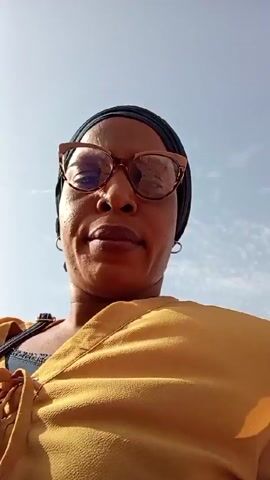 A person wearing glasses and a yellow shirt is facing the camera outside. The sky is visible in the background. The person is wearing a dark head covering. The person's face is in focus. The camera is pointed upwards at the person.  Sounds indicate the presence of vehicles.