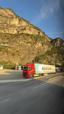 The video captures a view from inside a moving vehicle, likely a bus or a car, looking out a window. The scene is dominated by a towering, rocky mountain on the left, illuminated by the warm light of what appears to be the late afternoon or early morning sun. Green vegetation, including trees and shrubs, clings to the slopes of the mountain. A bright blue sky with wispy white clouds and a faint contrail stretches across the top of the frame.

In the foreground, a highway or a large road is visible. Several trucks are present on the road and parked in an adjacent area. The closest truck is a red semi-truck with a white trailer that has blue and white stripes and the text "BODNER" in large, dark letters. The truck is in motion, showing motion blur. Further down the road and in the parking area, other trucks are visible, some red, some white, and some with trailers of various colors, including green. A white van is also visible in the parking area. The lighting suggests a clear day. The perspective implies the viewer is traveling alongside these trucks.