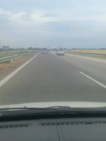 The video shows a view from the inside of a car driving on a highway. The highway has multiple lanes and is bordered by a guardrail on the left and fields on the right. Several cars and trucks are visible on the highway, moving in the same direction as the camera car. The sky is overcast.