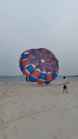 The video depicts a beach scene with a large, inflated parachute in the foreground. The parachute is primarily red, white, and blue, arranged in a circular pattern. Several individuals stand beneath the parachute, seemingly preparing for or engaging in parasailing activities. To the right of the parachute, a man in a white shirt and dark shorts stands alone, holding a long, green object. The beach is sandy with numerous footprints visible. The background includes the ocean, and distant land masses are visible on the horizon. The sky is overcast, suggesting a cloudy day. The overall setting suggests a recreational activity taking place at a beach resort.