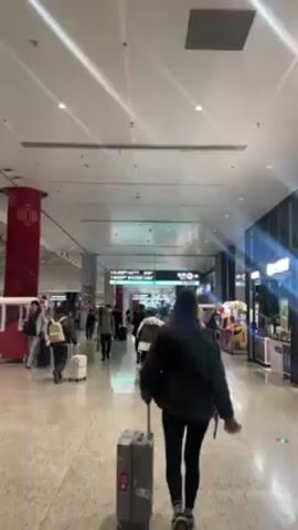 The video shows a person walking through an airport concourse, pulling a grey rolling suitcase behind them. The person is wearing a dark jacket, leggings, and a backpack. Other people walk in both directions, some also pulling luggage. There are small vendor carts on the right side of the corridor. Overhead signs in Chinese and English indicate directions. The concourse is brightly lit with a high ceiling.