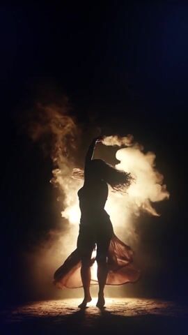 The media file depicts a silhouetted figure, appearing to be a woman, in motion against a dark background. She is suspended in the air, or perhaps falling, with her arms and legs extended in dramatic poses. A flowing garment, likely a dress or skirt, billows around her, catching the light and adding to the sense of movement and drama. Wisps of smoke or fog swirl around her, illuminated by a bright, unseen light source. The overall impression is one of an artistic performance, possibly dance or a theatrical scene, with a strong emphasis on silhouette, light, and shadow. There is no visible text within the media file. The setting appears to be a dark, possibly indoor space, with a textured vertical surface visible on the right side, which could be a wall or a backdrop. The time of day is indeterminate due to the artificial lighting and dark surroundings. The emotions conveyed are a mix of grace, power, and perhaps a hint of mystery or intensity.