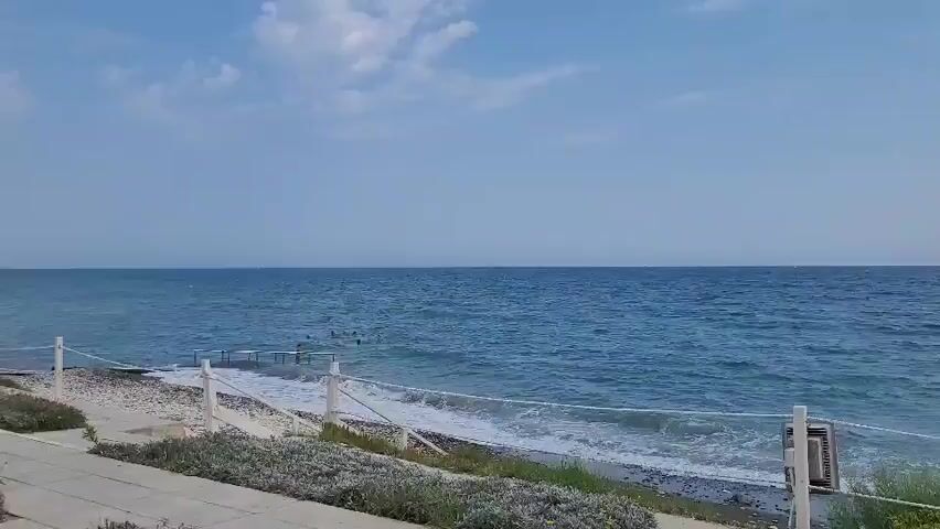 The video shows a view of the ocean from a beach. The water is blue and there are some waves. There are a few people in the water swimming. There is a white rope fence in the foreground. The sky is blue with some clouds. The sun is shining. The scene is calm and peaceful.