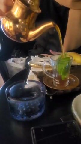 The video shows someone pouring steaming tea into a clear glass mug, which sits on a small, round, bronze saucer.  The mug already contains a brown liquid and a sprig of fresh mint. The tea is being poured from a golden, Aladdin-style teapot held above the mug. The steam from the tea obscures the person's face. In the foreground, a translucent blue glass ashtray is visible. Behind the mug, there is a golden-rimmed plate with a white napkin and a piece of fruit on it. Other plates and a cell phone are placed on the black table.