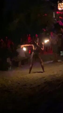The video shows a woman performing a fire dance on a beach at night. The woman, wearing dark clothing, stands on a sandy beach surrounded by an audience. She skillfully twirls two flaming torches, moving her arms and body in time with the music. The audience, a diverse group of people, stands or sits watching the performance, their silhouettes visible against the backdrop of the beach and some dimly lit structures. The scene is bathed in the orange glow of the fire, creating a lively and festive atmosphere.