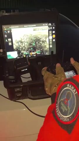 A person wearing a red jacket with a patch on the sleeve is operating a drone. The drone's control system is set up on a white surface, possibly the hood of a car. The control system includes a tablet displaying a thermal image from the drone, a radio, and a joystick. The tablet screen shows a grid overlay and a crosshair, suggesting the drone is being used for surveillance or search and rescue. The lighting in the scene is dim, indicating it may be nighttime or an indoor environment. The person's hands, wearing camouflage gloves, are visible on the joystick.