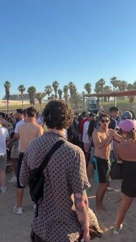The video shows a gathering of people on a sandy beach, likely at a party or event. Palm trees are visible in the background, along with a structure that appears to be a bar or stage area. Several individuals are standing, talking, and moving around. Some are shirtless, suggesting warm weather. The sun is visible, casting a warm glow on the scene, indicating it is likely daytime or late afternoon. The overall atmosphere appears casual and social.