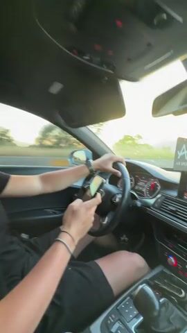A person is driving a car on a highway. The camera angle is from the driver's perspective, showing the dashboard and windshield. The sky is partly cloudy with a sunset visible in the distance. The person is holding a mobile phone in their left hand while their right hand is on the steering wheel. The car's infotainment system displays music information. There is an air freshener hanging from the rearview mirror. The environment is a highway with mountains in the background.
