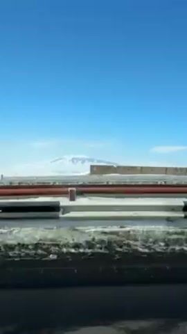 The video shows a view from a moving vehicle on a highway. A concrete barrier runs along the edge of the road. Beyond the barrier is a snowy field, and in the distance, a snow-capped mountain is visible under a clear blue sky. The road is asphalt and shows signs of wear.