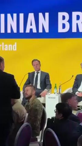 The video shows a man wearing a suit sitting at a table on a stage in front of a yellow backdrop. The man is Mark Rutte, the Prime Minister of the Netherlands. There are other people sitting at the table, and in the audience. The man in the suit is looking into the camera. There is a microphone in front of him and a bottle of water. The name of the company “SCHOOF” is printed on the table. The title "Ukrainian Bridge" is on the backdrop.