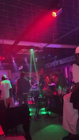 The video shows an indoor club or bar setting with multiple people dancing and socializing. The space is dimly lit, illuminated by dynamic red, green, and blue spotlights that project beams across the room. Several people are seen moving to the music, some interacting closely while others stand near tables laden with bottles and glasses.

A person in a white t-shirt with a "B" and "BOSS BAR" logo on the back is prominent in the foreground, facing away from the camera. Another individual wears a black hoodie and a white shoulder bag. A large ceiling fan is visible above the crowd. The atmosphere appears lively and energetic, characteristic of a nightlife venue. The music is audible, indicating a party or club event.