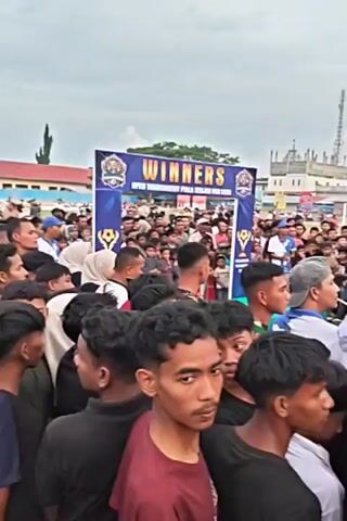 The video shows a large crowd of people gathered outdoors, possibly at an event or competition. In the background, there are banners that say "WINNERS" and "OPEN CHAMPIONSHIP." Several people are wearing blue shirts and caps, while others are in casual attire. Some individuals appear to be speaking or addressing the crowd, while others are observing. There are also tents set up, suggesting a temporary event space. The overall atmosphere seems lively and engaging.