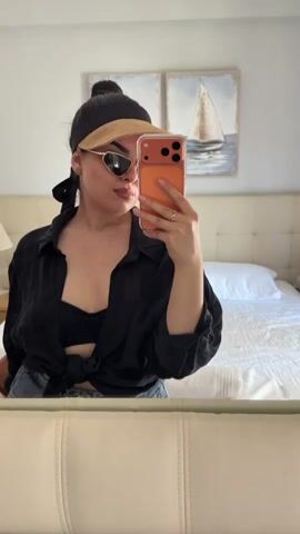 A woman in a black top and jeans takes a selfie in a bedroom. She is wearing sunglasses and a baseball cap. Behind her, there is a bed with white sheets and pillows, and two paintings of sailboats on the wall.