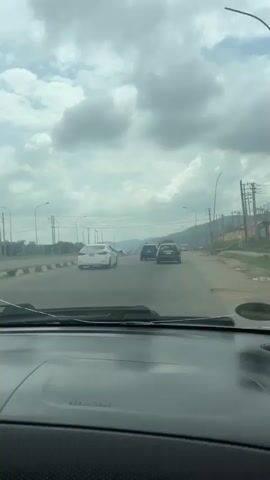 The video shows a view from inside a car driving on a road. The sky is overcast with clouds. There are several cars on the road ahead, including a white sedan, a black SUV, and a green car. The road appears to be a multi-lane highway with streetlights and utility poles lining the sides. There are some buildings and vegetation visible in the distance, with hills in the background. The camera is positioned behind the dashboard, showing the windshield and the front of the car.