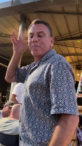 A man wearing a patterned short-sleeved shirt is speaking animatedly, gesturing with his left hand. He is outdoors under a covered structure with exposed beams and string lights. In the background, several people are visible. A woman in the foreground is holding up a white object and appears to be singing or speaking. Further back, a group of people are seated and standing, some appearing to be clapping or engaged in conversation. The overall atmosphere seems to be a casual social gathering.